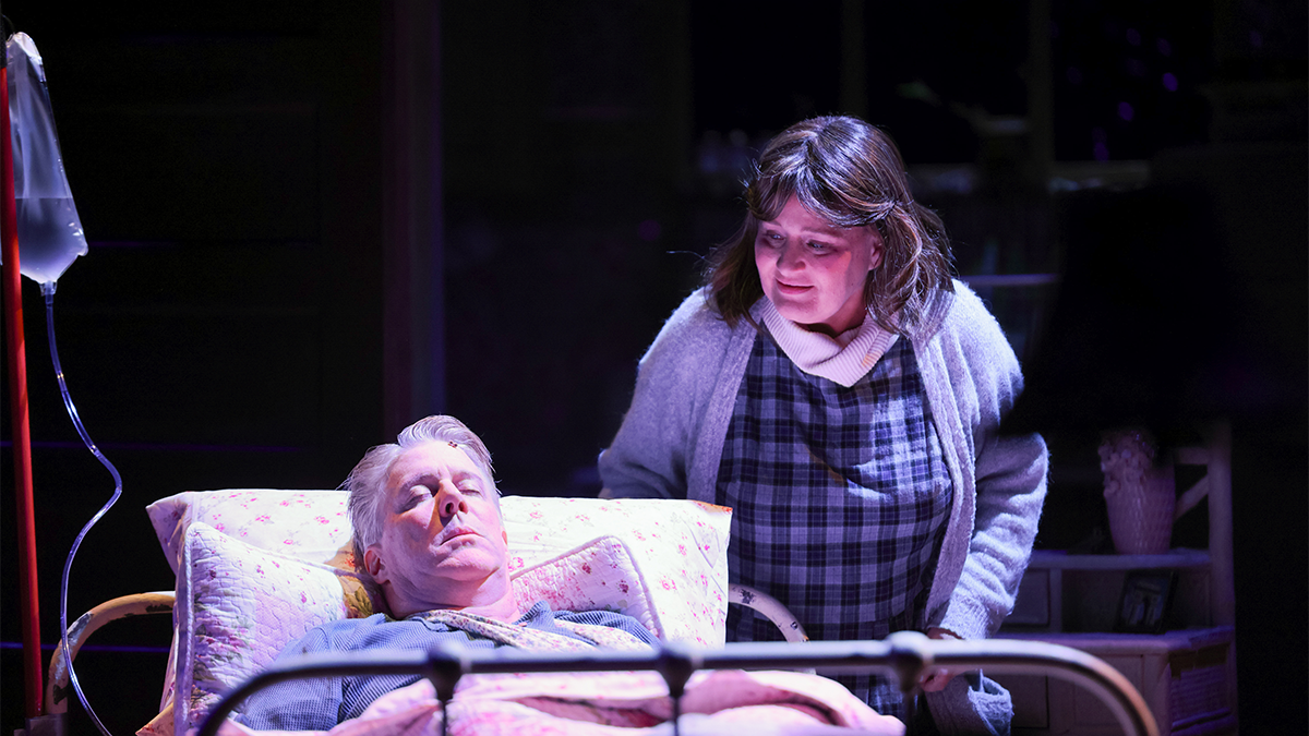A woman stands over a wounded man in bed. Both on stage.